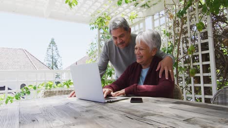 Happy-senior-mixed-race-couple-sitting-in-garden-using-laptop