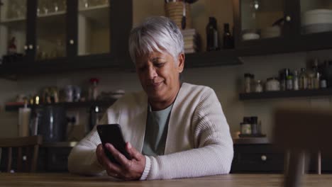 Lächelnde-ältere-Frau-Mit-Gemischter-Abstammung,-Die-In-Der-Küche-Ihr-Smartphone-Benutzt