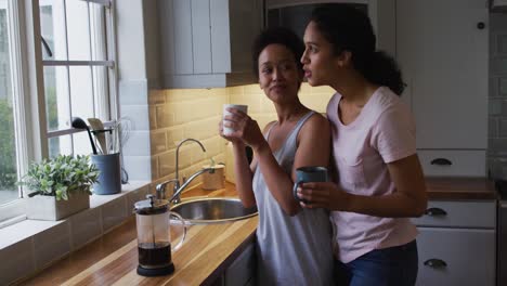 Mixed-race-lesbian-couple-hugging-and-drinking-coffee-in-kitchen
