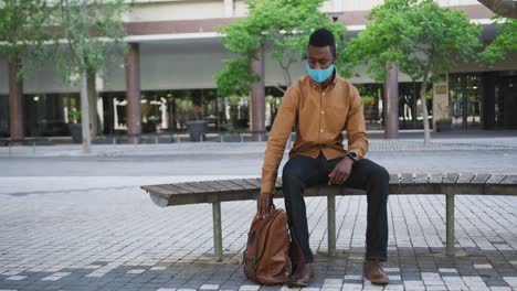 African-american-businessman-wearing-face-mask-sitting-on-bench
