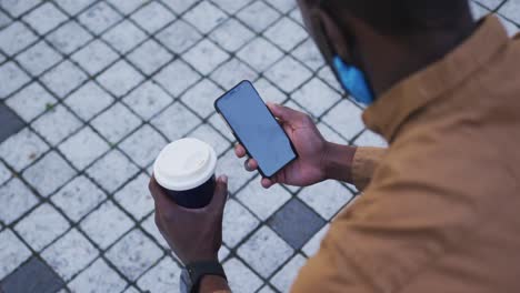 Hombre-De-Negocios-Afroamericano-Con-Mascarilla-Usando-Un-Teléfono-Inteligente-Sosteniendo-Café