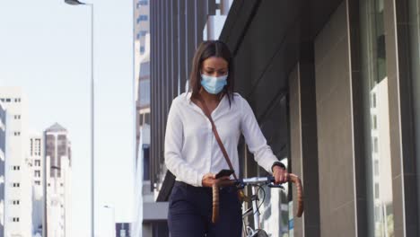 African-american-woman-wearing-face-mask-using-smartphone-wheeling-bicycle