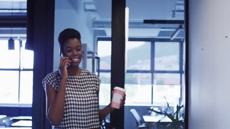 African-american-businesswoman-talking-on-smartphone-holding-coffee-walking-in-office-corridor