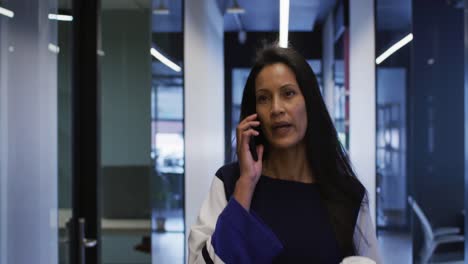 Mixed-race-businesswoman-talking-on-smartphone-holding-coffee-walking-in-office-corridor