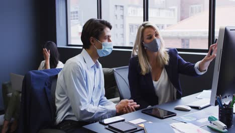 Diverse-business-people-wearing-face-masks-using-computer-in-office