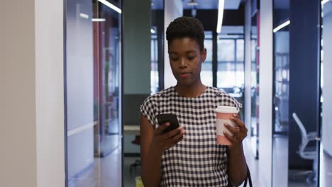 African-american-businesswoman-using-smartphone-holding-coffee-walking-in-office-corridor