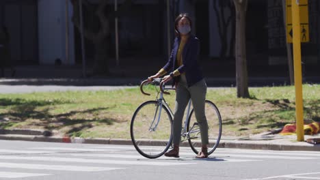 African-american-woman-wearing-face-mask-wheeling-bicycle-crossing-road