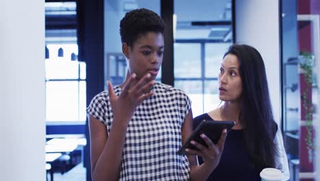 Diverse-businesswomen-talking-using-digital-tablet-walking-through-office