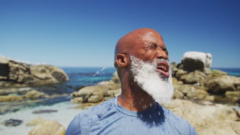 Älterer-Afroamerikanischer-Mann-Trainiert,-Indem-Er-Auf-Felsen-Am-Meer-Wasser-Auf-Sein-Gesicht-Schüttet