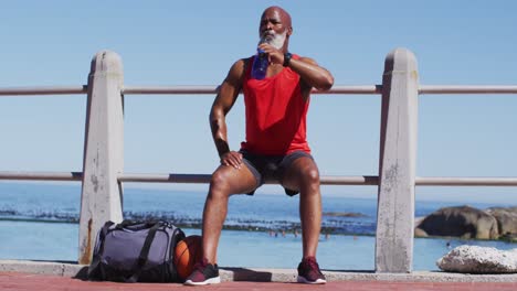 Älterer-Afroamerikanischer-Mann-Trinkt-Wasser,-Während-Er-Auf-Dem-Geländer-In-Der-Nähe-Des-Strandes-Sitzt