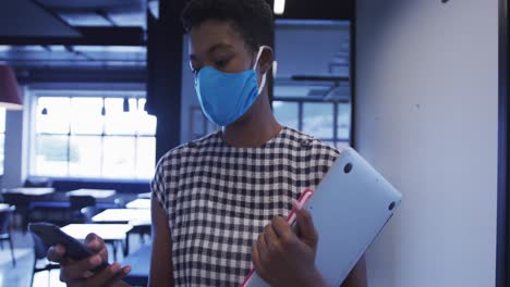 African-american-businesswoman-wearing-face-mask-holding-paperwork-using-smartphone-walking-in-offic