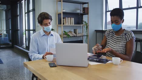 Diverse-business-collegues-wearing-face-masks-sitting-using-laptop-going-through-paperwork-in-office