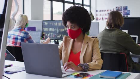 Mixed-race-businesswoman-wearing-face-mask-sitting-using-a-laptop-in-modern-office