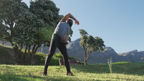 Senior-african-american-man-wearing-face-mask-exercising-stretching-in-countryside