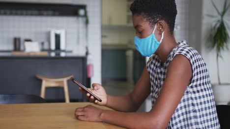 African-american-businesswoman-wearing-face-mask-sitting-in-workplace-kitchen-using-smartphone