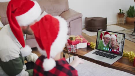 Caucasian-man-with-son-wearing-santa-hats-on-laptop-video-chat-during-christmas-at-home