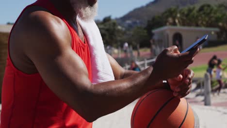 Senior-african-american-man-with-basketball-using-smartphone-near-the-beach