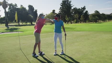 Two-caucasian-women-playing-golf-wearing-face-masks-greeting-each-other-with-elbows