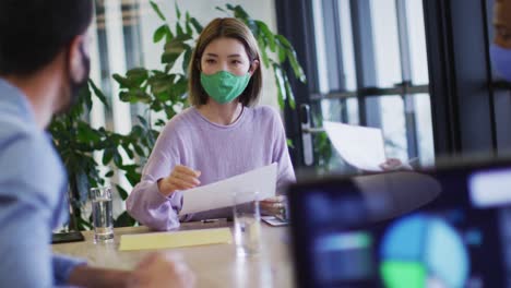 Diverse-business-people-wearing-face-masks-sitting-using-laptops-going-through-paperwork-in-office