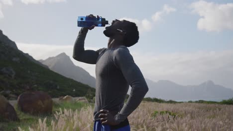 Hombre-Afroamericano-Bebiendo-Agua-De-Una-Botella-Mientras-Camina-Por-Las-Montañas