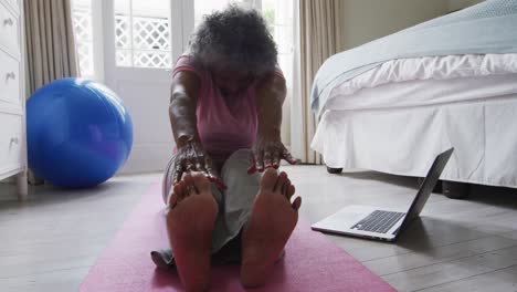 Senior-african-american-woman-performing-stretching-exercise-while-looking-at-laptop-at-home
