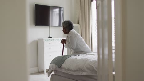 Senior-african-american-woman-holding-walking-stick-sitting-on-the-bed-at-home