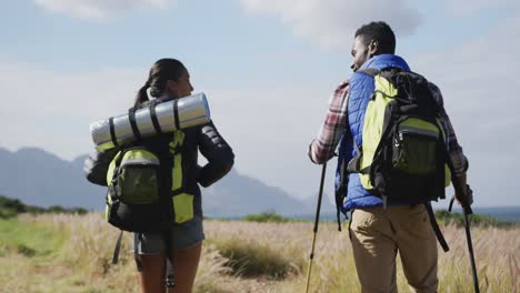 Diverse-couple-wearing-backpacks-using-nordic-walking-poles-hiking-in-countryside
