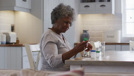 Mujer-Afroamericana-Mayor-Mirando-Un-Contenedor-De-Medicamentos-Vacío-Mientras-Está-Sentada-En-Casa
