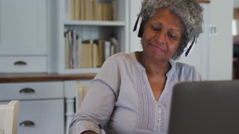 Ältere-Afroamerikanische-Frau-Trägt-Ein-Telefon-Headset-Und-Macht-Sich-Notizen,-Während-Sie-Zu-Hause-Einen-Laptop-Benutzt