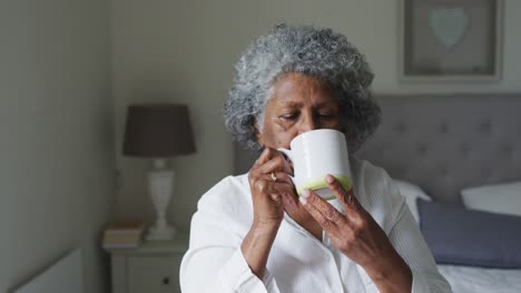 Ältere-Afroamerikanische-Frau-Trinkt-Kaffee,-Während-Sie-Zu-Hause-Auf-Dem-Bett-Sitzt