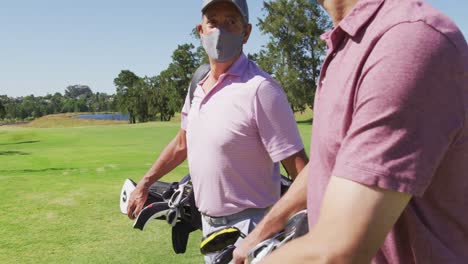 Two-caucasian-senior-man-wearing-face-masks-walking-with-their-golf-bags-at-golf-course