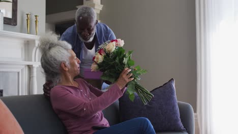 Afroamerikanischer-älterer-Mann,-Der-Seiner-Frau-Zu-Hause-Eine-Geschenkbox-Und-Einen-Blumenstrauß-Schenkt