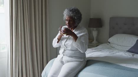 Ältere-Afroamerikanische-Frau-Trinkt-Kaffee,-Während-Sie-Zu-Hause-Auf-Dem-Bett-Sitzt