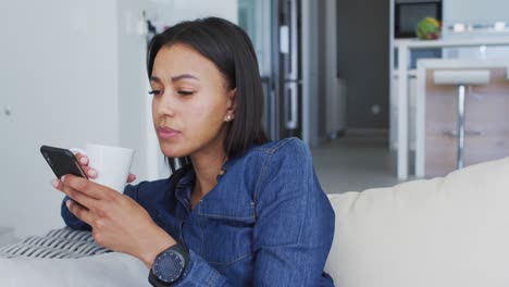 Mixed-race-woman-sitting-on-couch-using-smartphone-drinking-cup-of-coffee