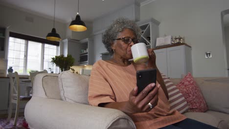 Ältere-Afroamerikanische-Frau,-Die-Ihr-Smartphone-Benutzt,-Während-Sie-Zu-Hause-Auf-Der-Couch-Kaffee-Trinkt
