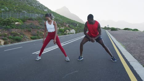 Diverse-fit-couple-exercising-stretching-on-a-country-road-near-mountains