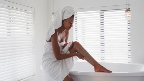 African-american-woman-applying-moisturizer-on-her-legs-in-the-bathroom-at-home