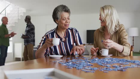 African-american-and-caucasian-senior-women-sitting-by-table-doing-puzzles-drinking-tea