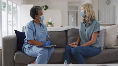 Senior-african-american-female-doctor-talking-with-female-patient-at-home-both-wearing-face-masks