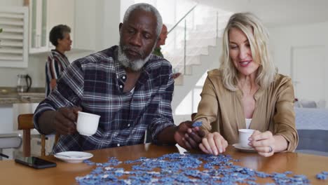 African-american-senior-man-and-caucasian-senior-woman-sitting-by-table-doing-puzzles-drinking-tea