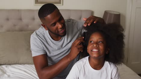 Afroamerikanischer-Vater-Bürstet-Die-Haare-Seiner-Tochter,-Während-Er-Zu-Hause-Auf-Dem-Bett-Sitzt