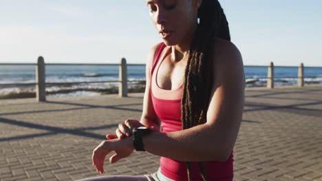 African-american-woman-in-sportswear-checking-smartwatch-on-promenade-by-the-sea