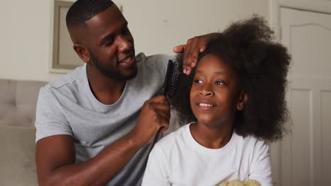 Afroamerikanischer-Vater-Bürstet-Die-Haare-Seiner-Tochter,-Während-Er-Zu-Hause-Auf-Dem-Bett-Sitzt