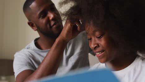 Afroamerikanischer-Vater,-Der-Seiner-Tochter-Die-Haare-Bindet,-Während-Er-Zu-Hause-Auf-Dem-Bett-Sitzt