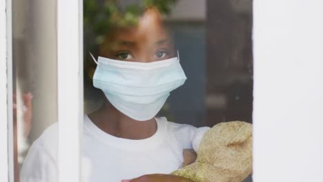 African-american-girl-wearing-face-mask-holding-teddy-bear-looking-through-window