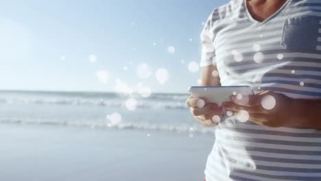 Animation-of-multiple-white-spots-over-midsection-of-woman-using-smartphone-by-seaside
