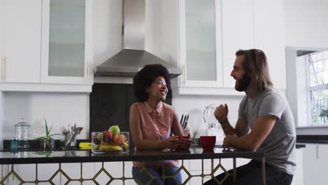 Mixed-race-couple-holding-coffee-cup-talking-to-each-other-in-the-kitchen-at-home