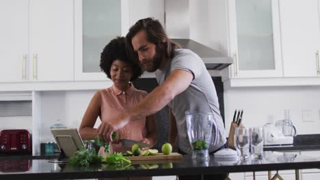 Mixed-race-couple-preparing-vegetable-smoothie-while-looking-at-digital-tablet-in-the-kitchen-at-hom