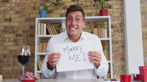 Caucasian-man-making-video-call-holding-handwritten-sign-making-marriage-proposal-and-celebrating-ac