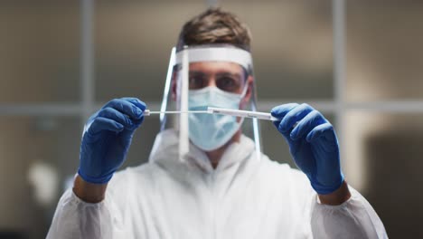 Caucasian-male-medical-worker-wearing-protective-clothing-and-face-shield-inspecting-dna-swab-in-lab
